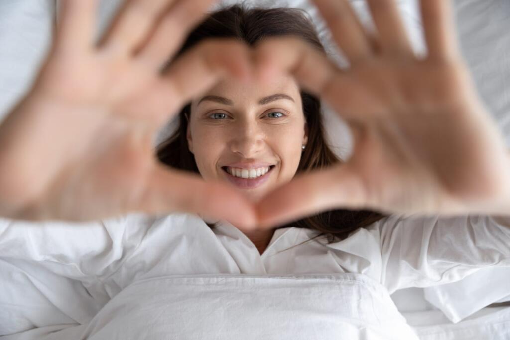 A woman in bed, getting ready to go to sleep and making a heart symbol with her hands.
