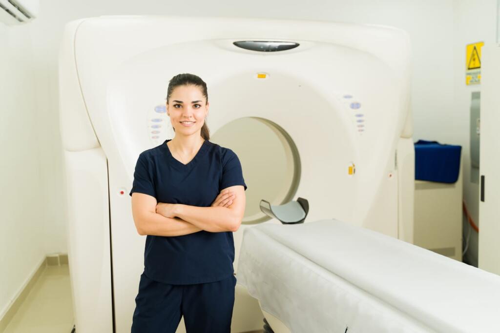 A woman in blue scrubs is smiling with her arms crossed as she poses for a photo in front of a CT scanner. 