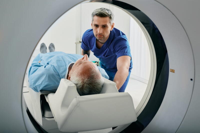 A male nurse is assisting an elderly male patient into a CT scanner to screen for lung cancer.