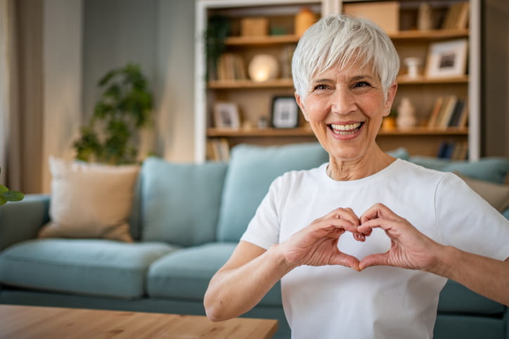 An elderly woman making a heart with her hands and holding it in front of her chest. 