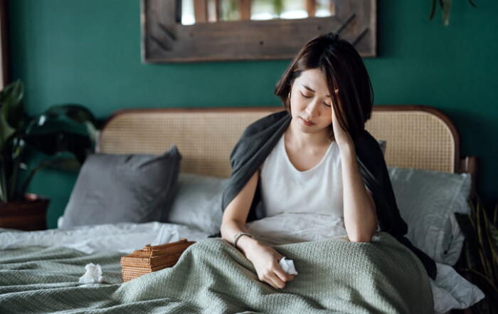 A Woman Sitting In Bed With Her Head In Her Hand As Her Chronic Stress Affects Her Health.