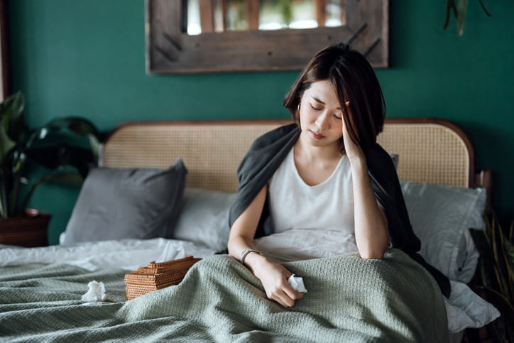 A Woman Sitting In Bed With Her Head In Her Hand As Her Chronic Stress Affects Her Health. 