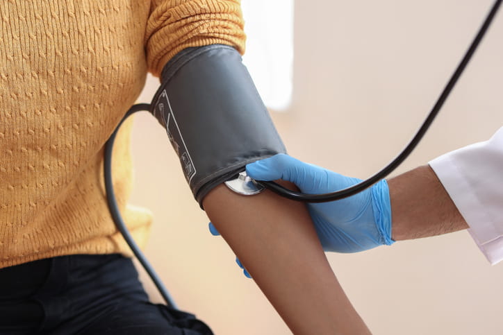 A Close-Up Of A Healthcare Provider'S Gloved Hand Using A Stethoscope And Blood Pressure Cuff To Check A Patient'S Arm. The Patient Is Wearing A Yellow Sweater.